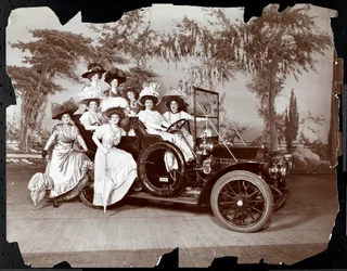 1906年の「ヴァンダービルトカップ」の公演であると思われる、景観を背景にしたステージ上の自動車内およびその周辺でポーズをとった8人の衣装を着た女性